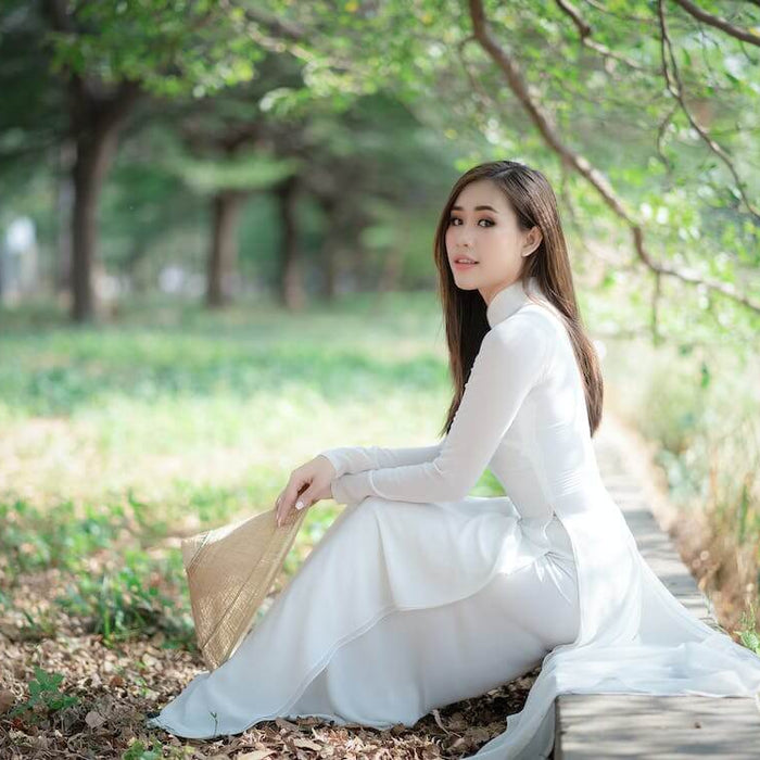 Woman in a vintage-inspired white dress sitting outdoors in a serene, leafy park.