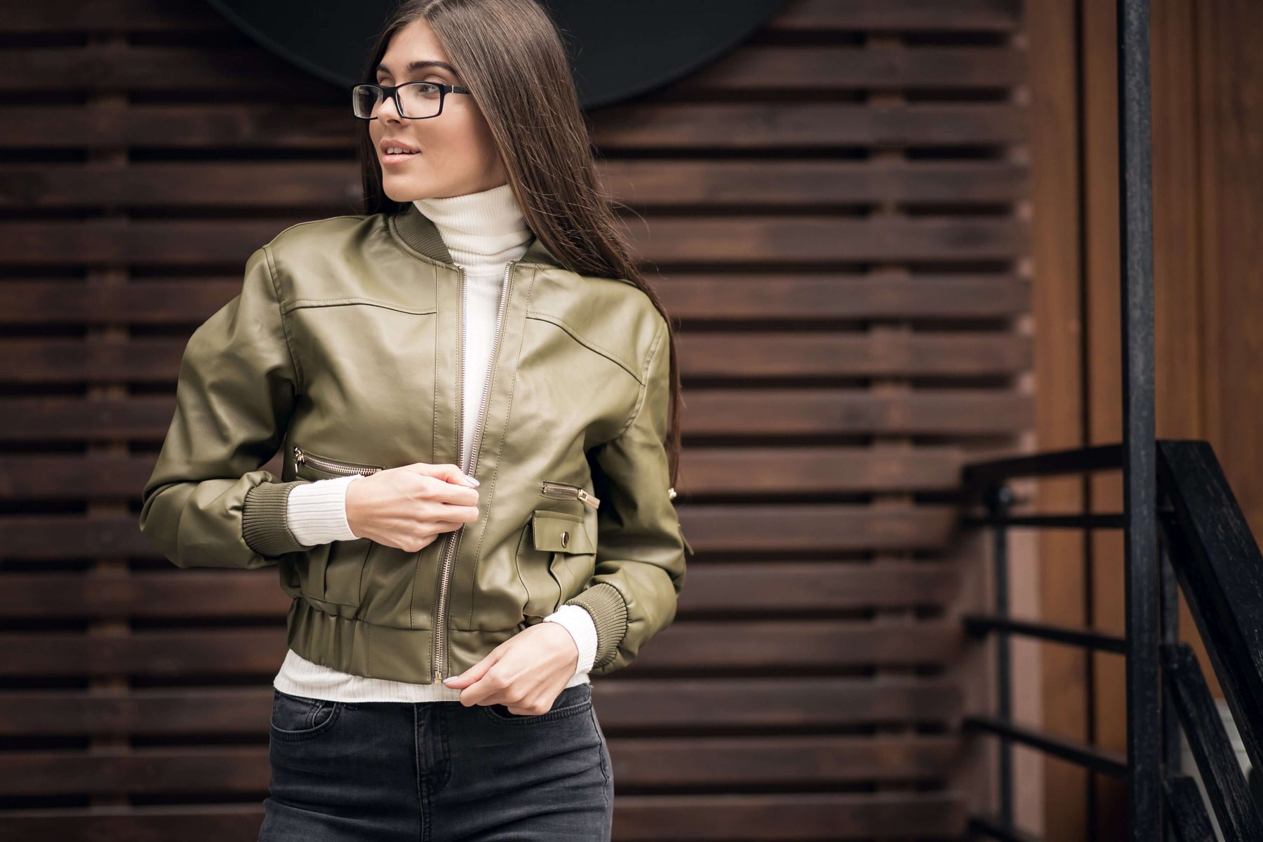 Woman wearing a stylish green leather bomber jacket, paired with a white turtleneck, standing in front of a wooden backdrop.