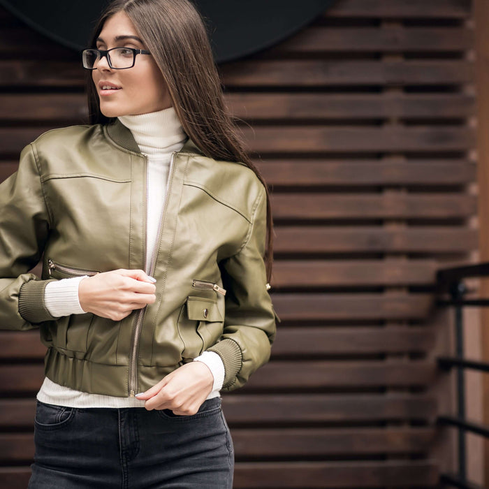 Woman wearing a stylish green leather bomber jacket, paired with a white turtleneck, standing in front of a wooden backdrop.